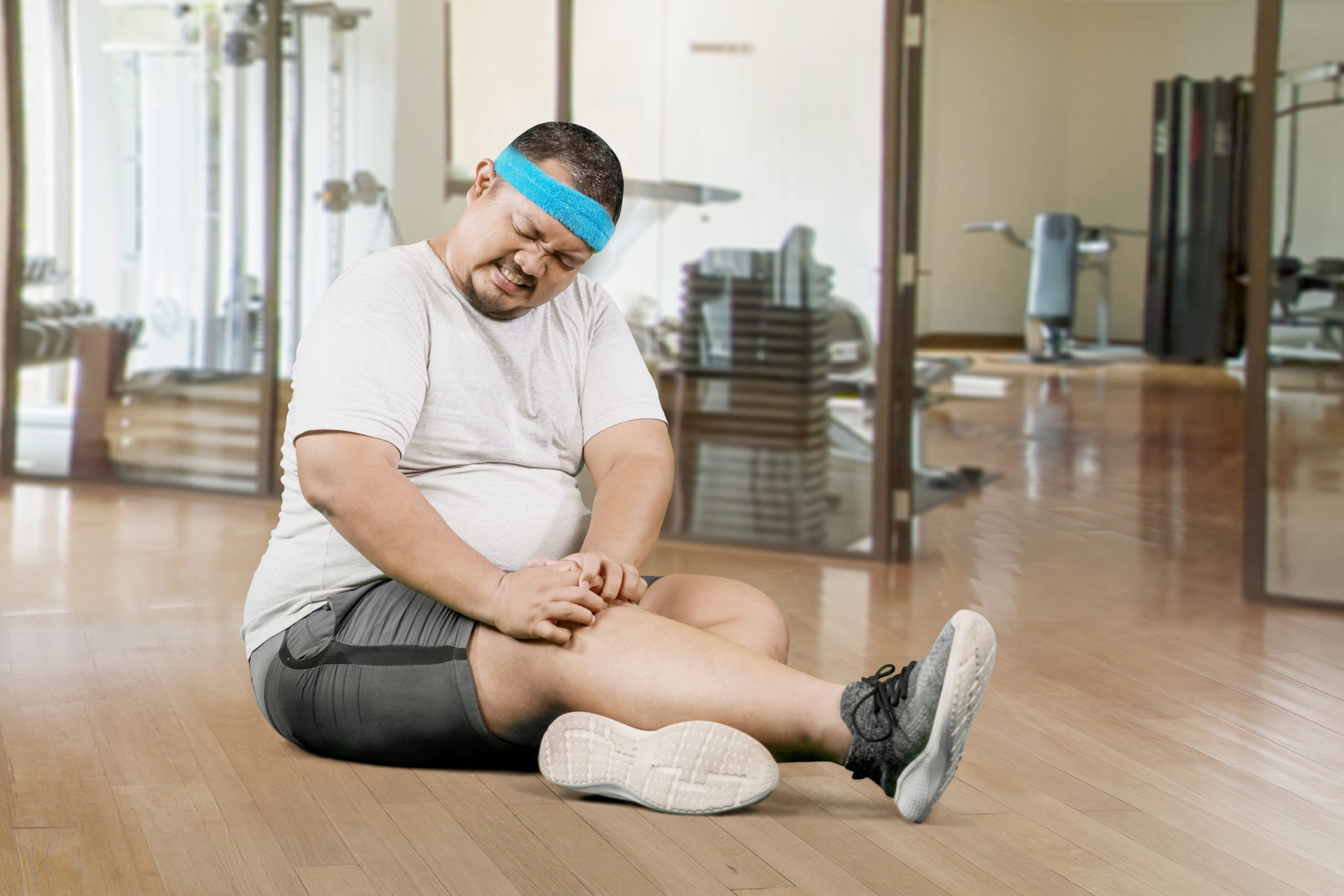 Portrait of young plus size overweight man holding a knee suffering from an injury in a gym