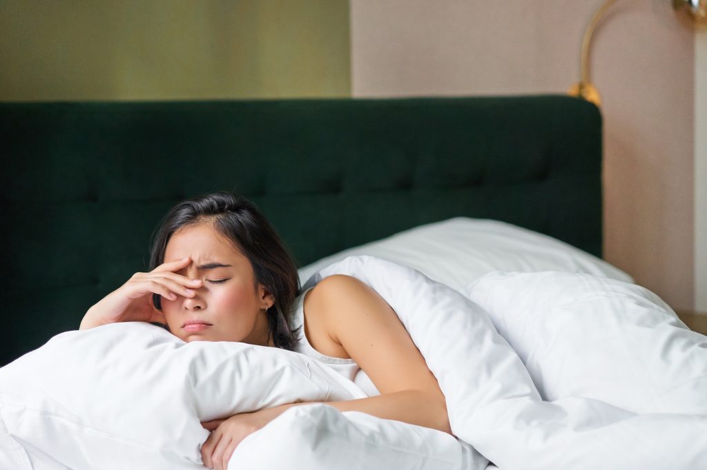 Sad and distressed young korean woman lying in her bed with pillow and warm duvet, feels uneasy, frowning and grimacing